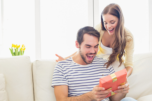 Pretty woman surprising her boyfriend with gift at home in the living room